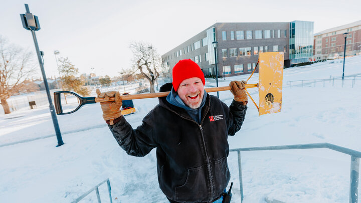 Kevin smiles for a photo while he holds a snow shovel across his shoulders
